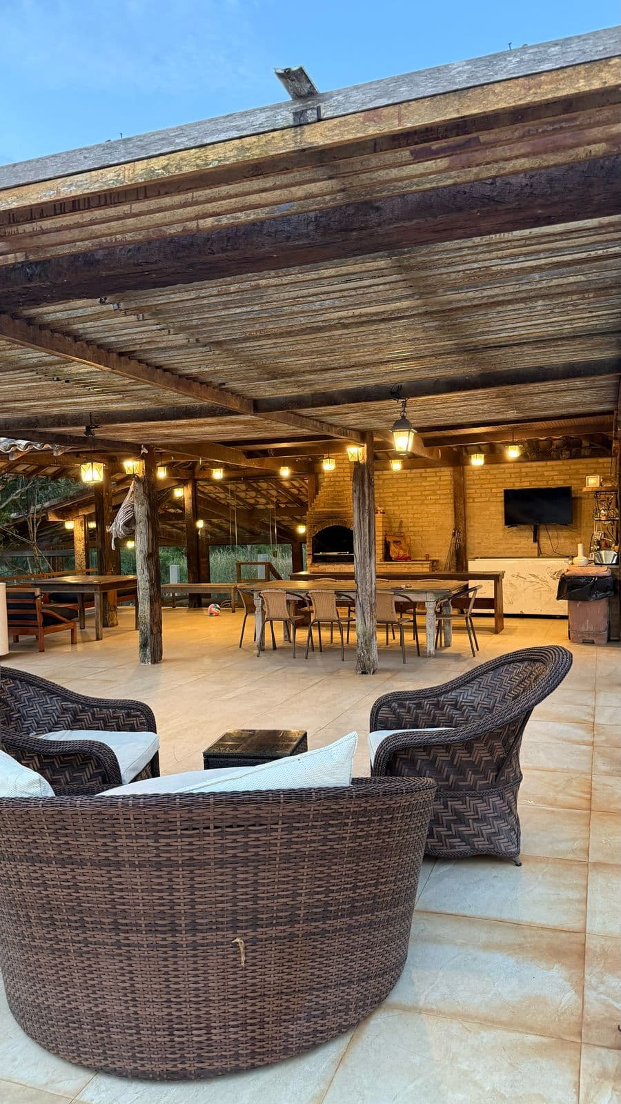Área de convivência ao entardecer, com poltronas, mesa, forno a lenha e luzes acesas